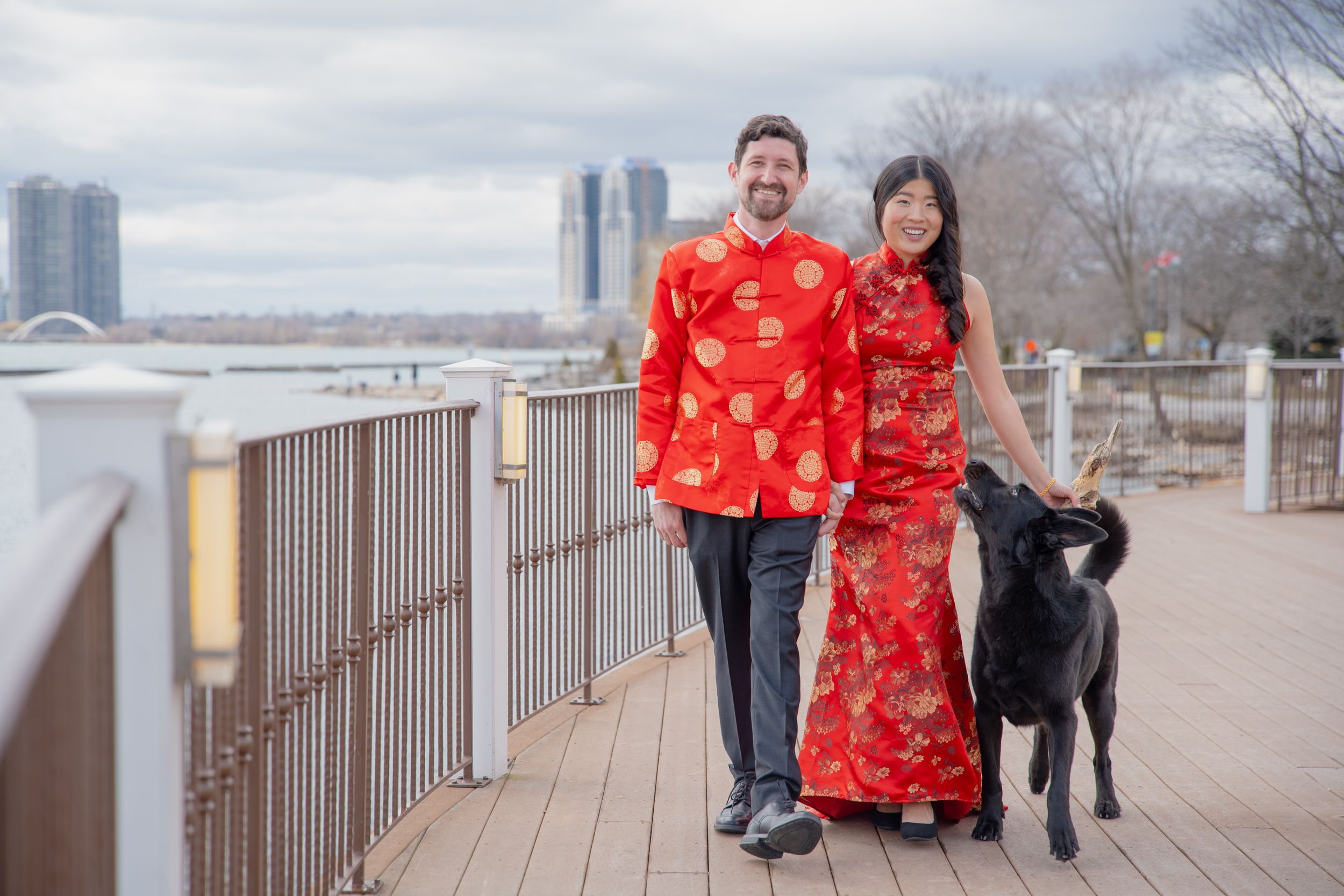 Couple in matching red Chinese outfits posing with black dog by waterfront.