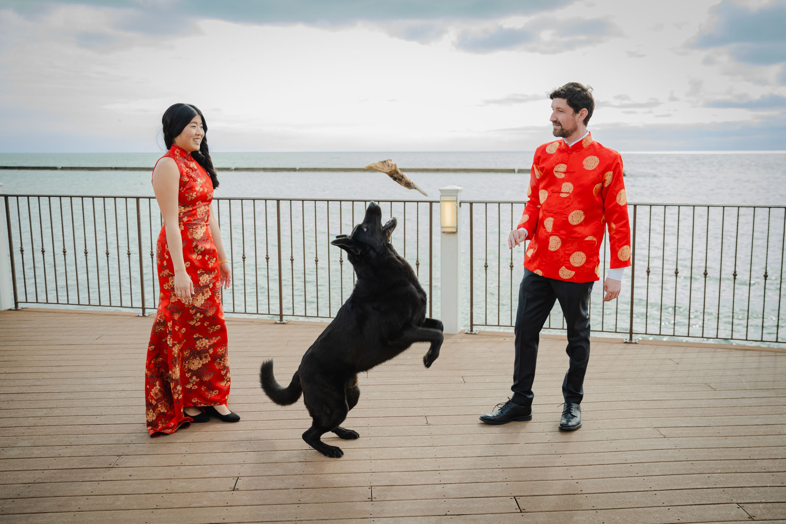Couple in red Chinese traditional outfits playing with black dog on waterfront boardwalk.