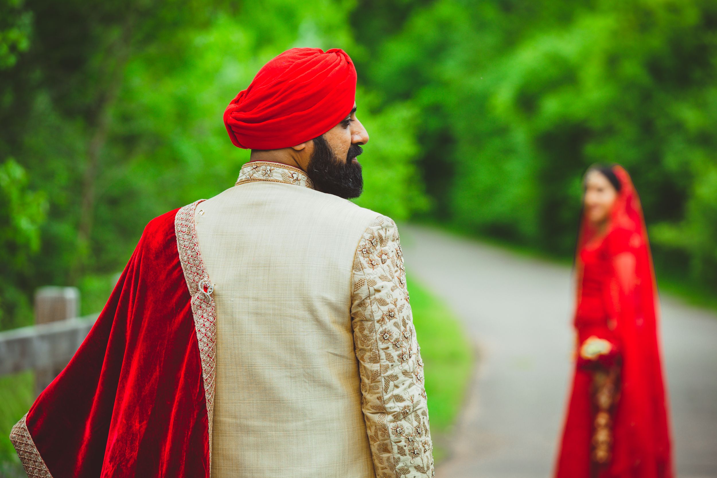 Traditional Sikh groom in red turban and velvet shawl standing on garden path.