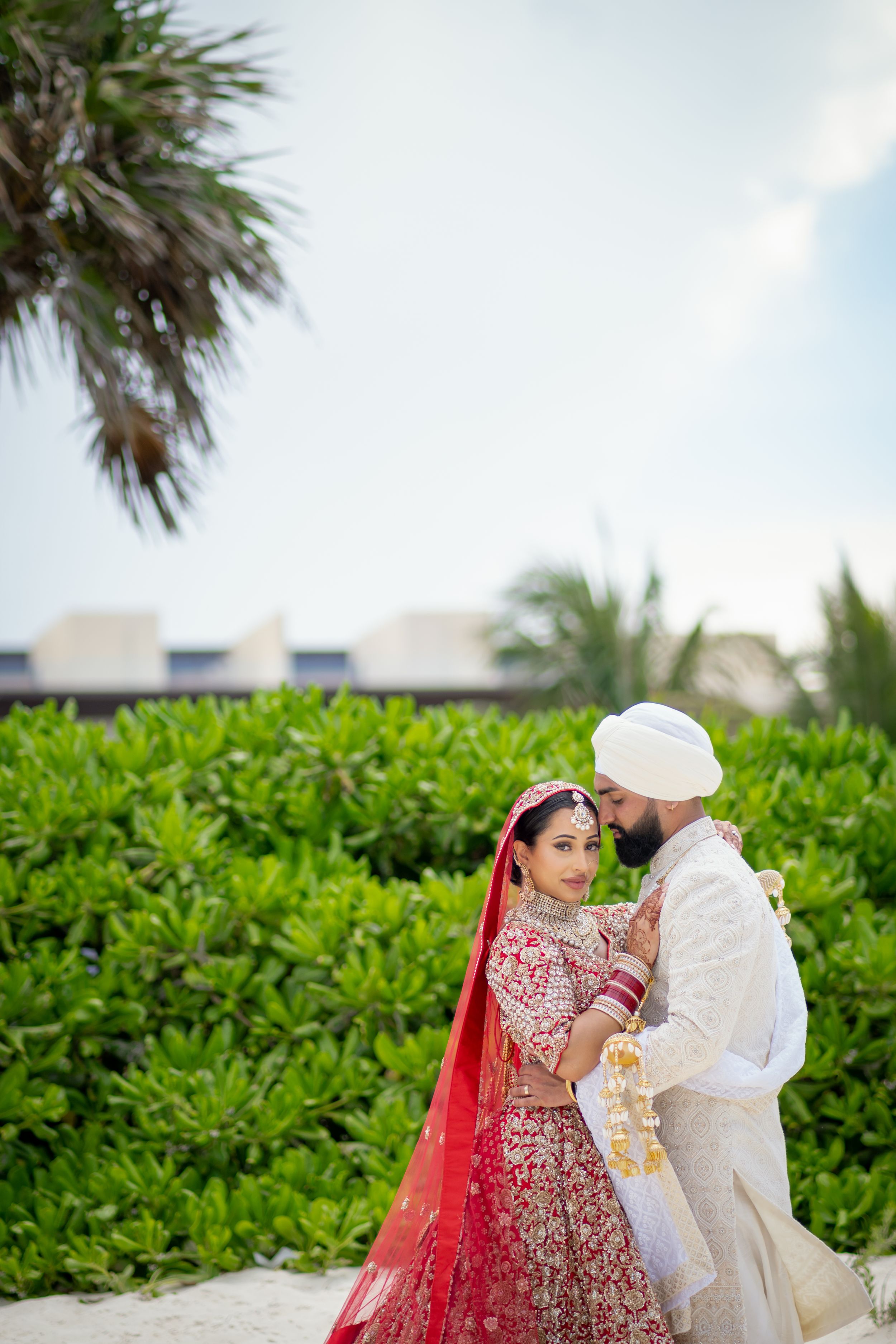 Couple in traditional Indian wedding attire poses against tropical garden backdrop.