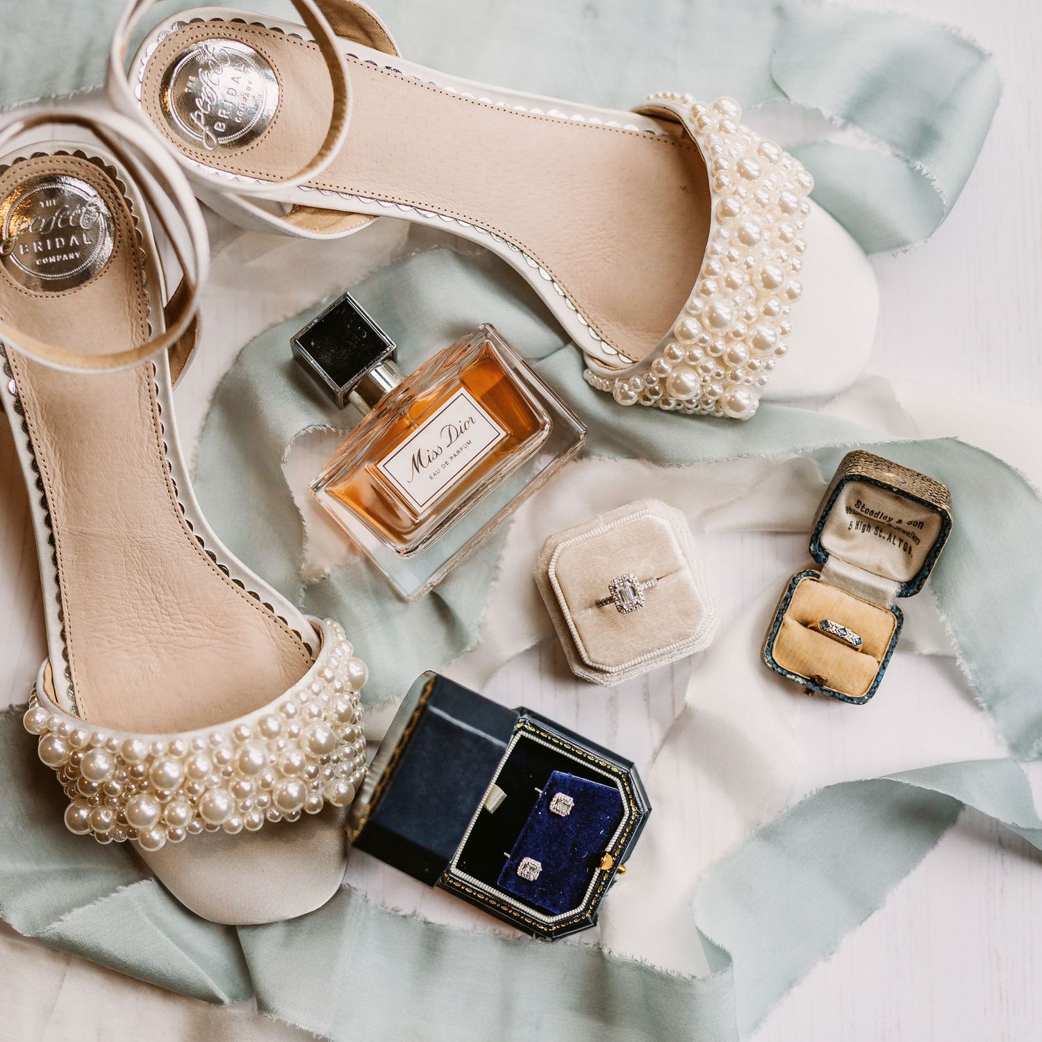 Elegant bridal accessories including pearl shoes, perfume, and jewelry boxes arranged on light fabric.