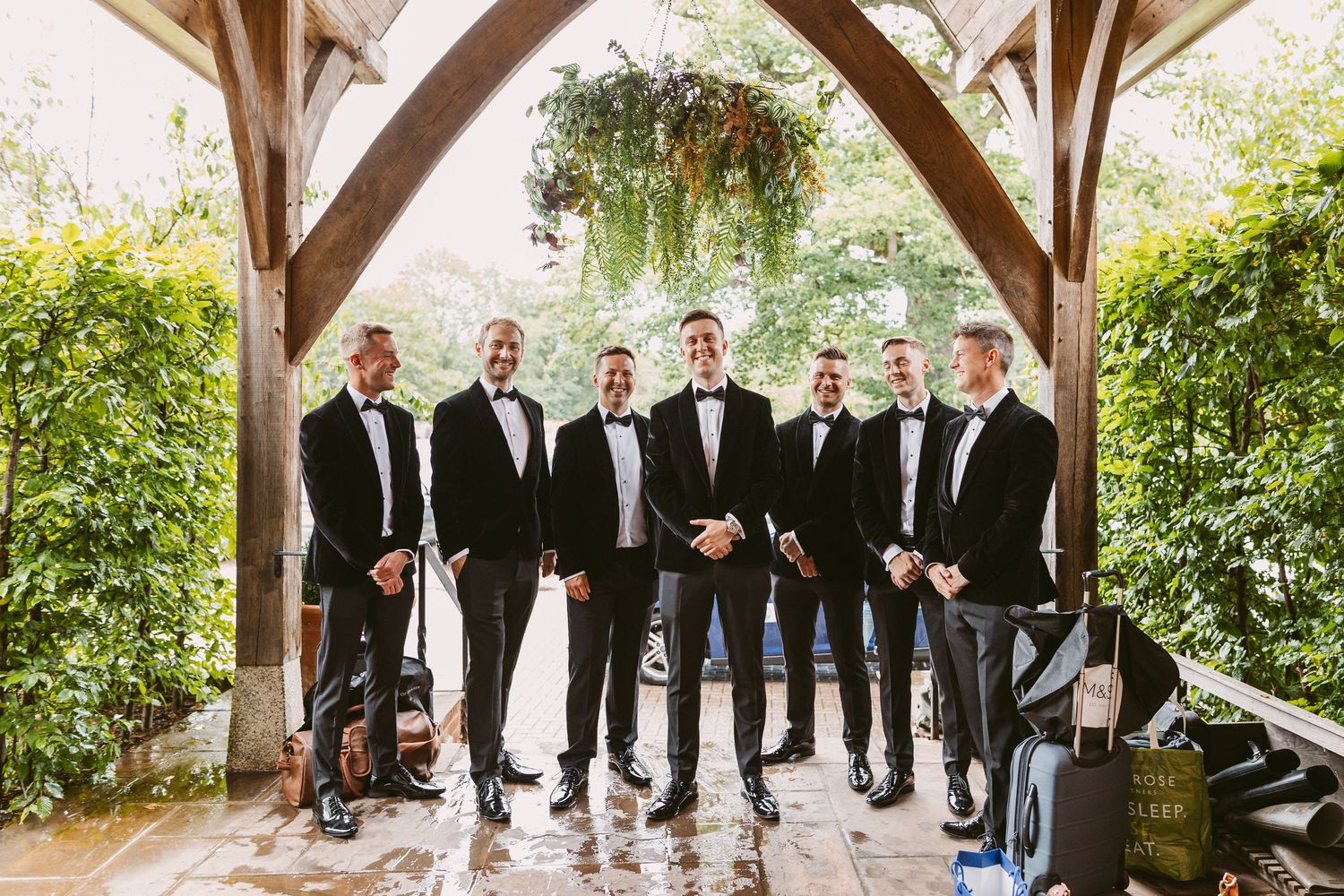 Formal group portrait of groomsmen in black tuxedos under rustic wooden archway.