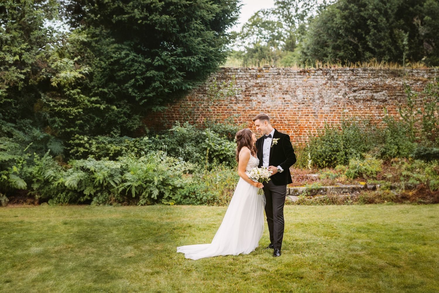 Couple posing in garden setting with rustic brick wall and greenery backdrop.