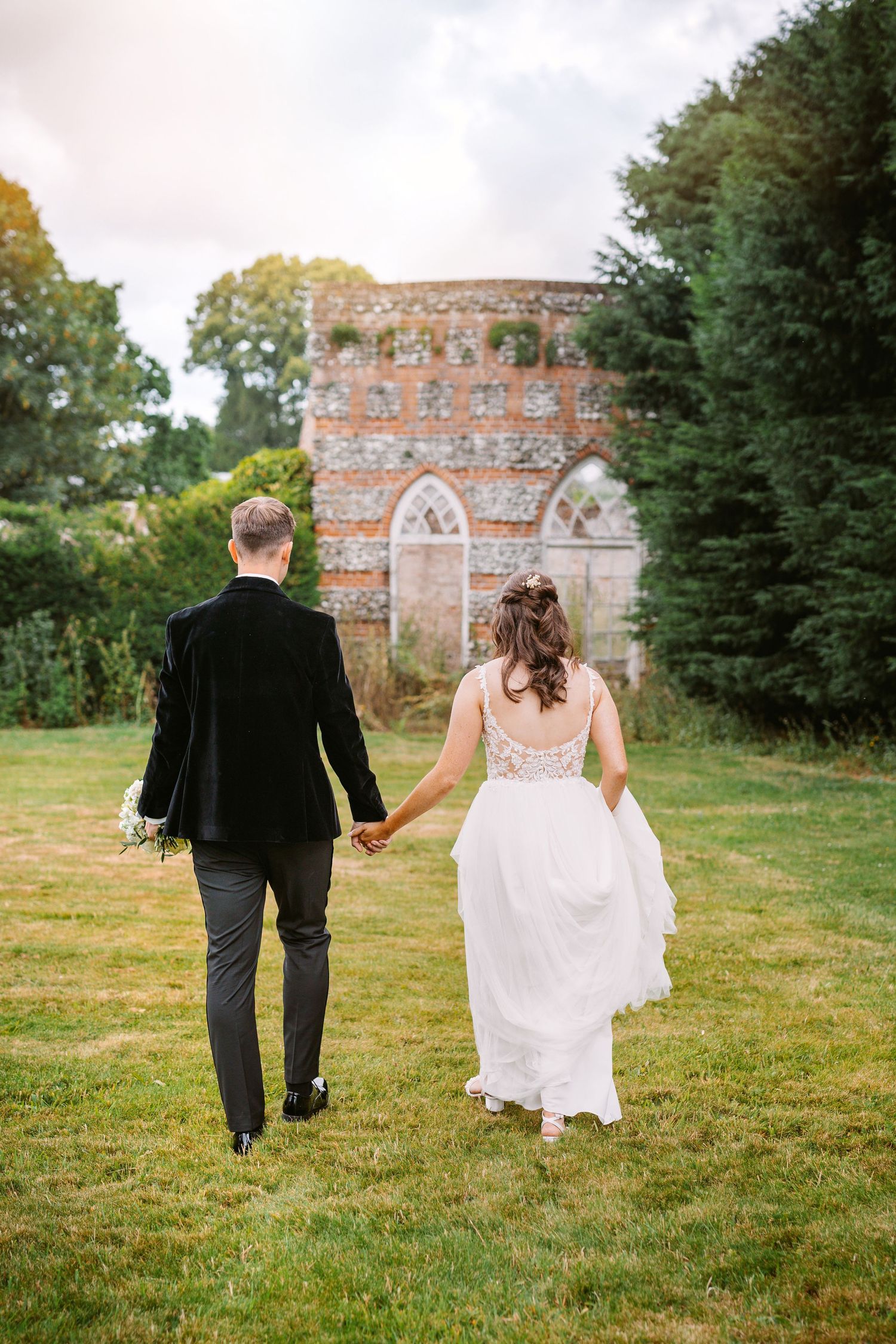 Couple walking hand in hand toward stone castle structure in garden setting.