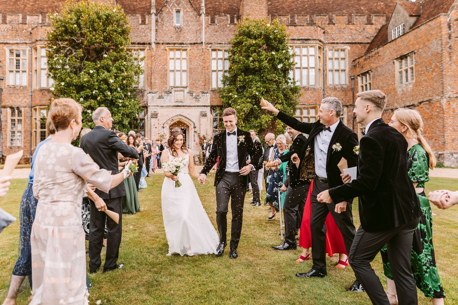 Wedding guests toss confetti at a celebration on the lawn of a historic brick manor house.