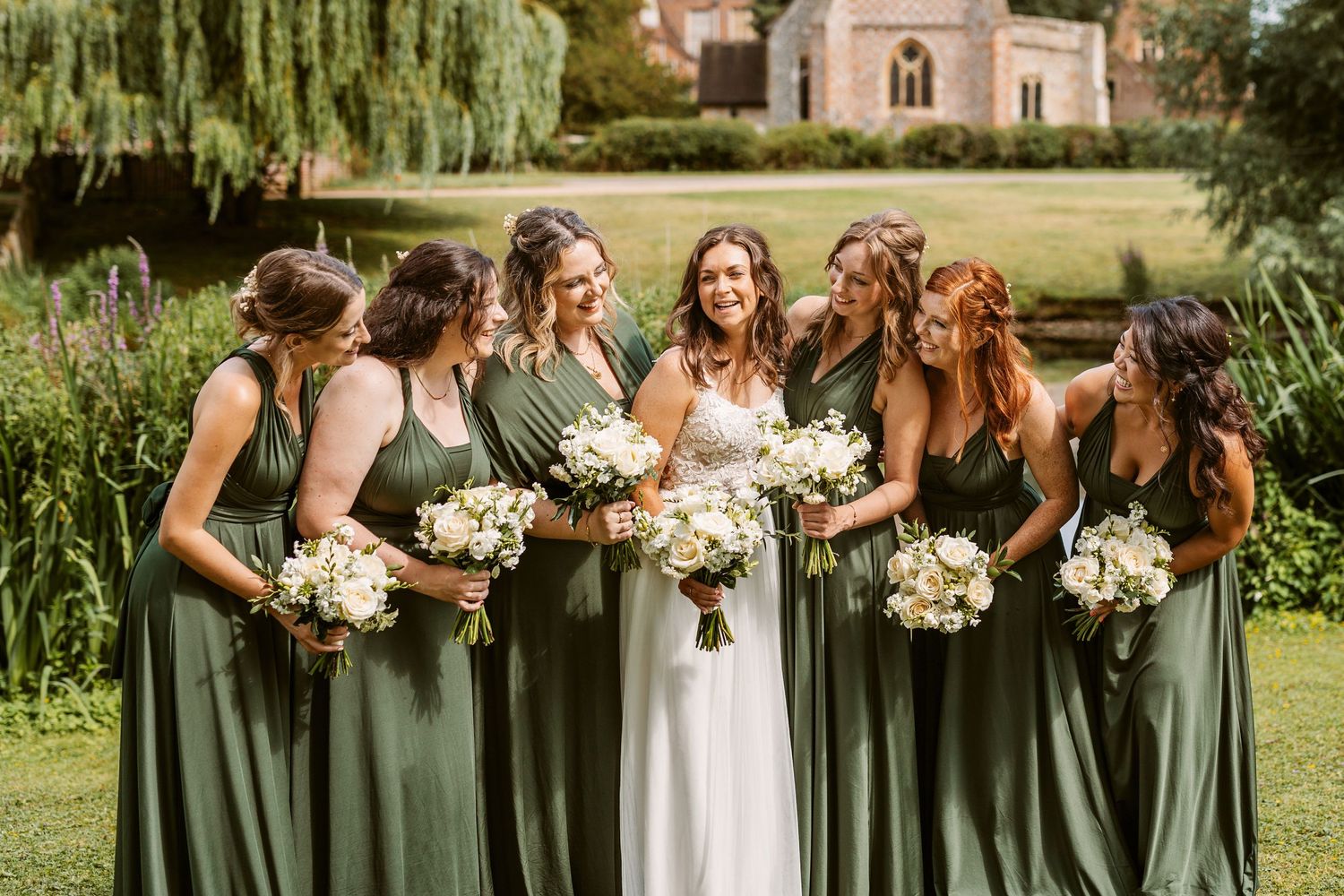 Wedding party in sage green dresses poses with bouquets on a sunny garden lawn.