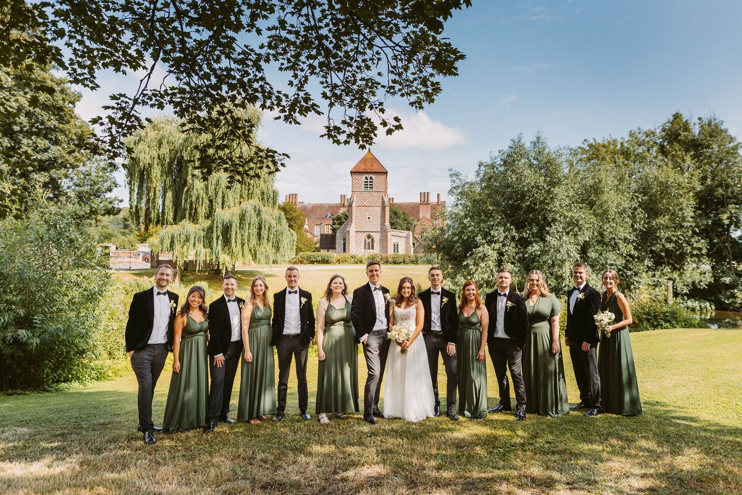 Wedding party in formal attire poses on lawn with historic building and clock tower in background.