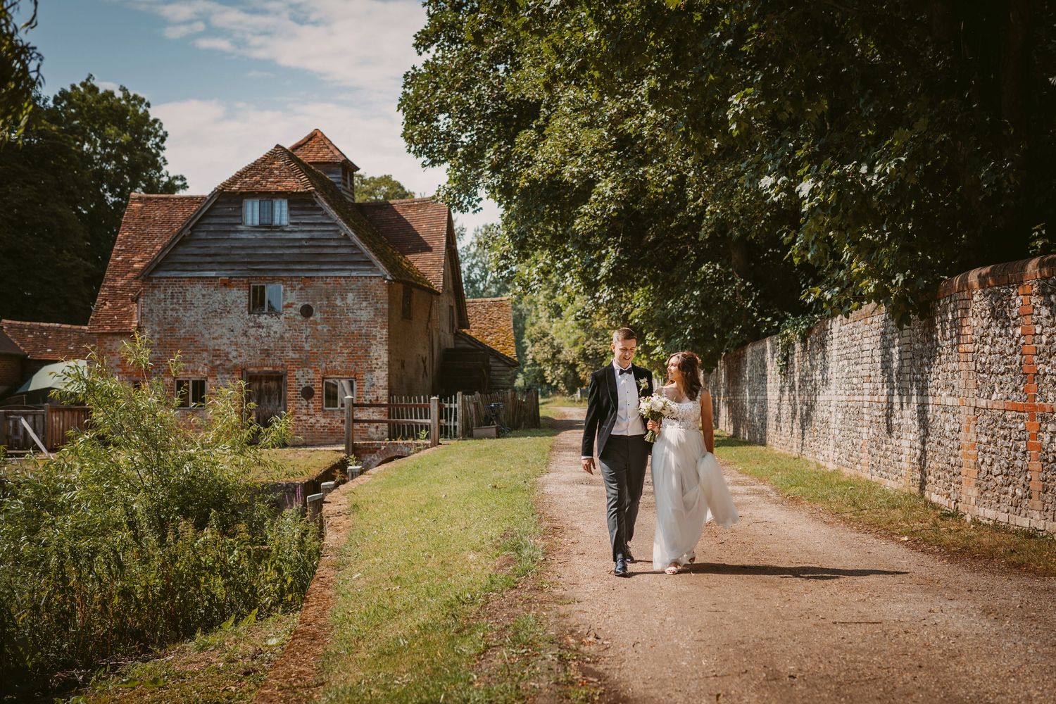 Couple walks along rustic path beside old brick wall and historic farm buildings.