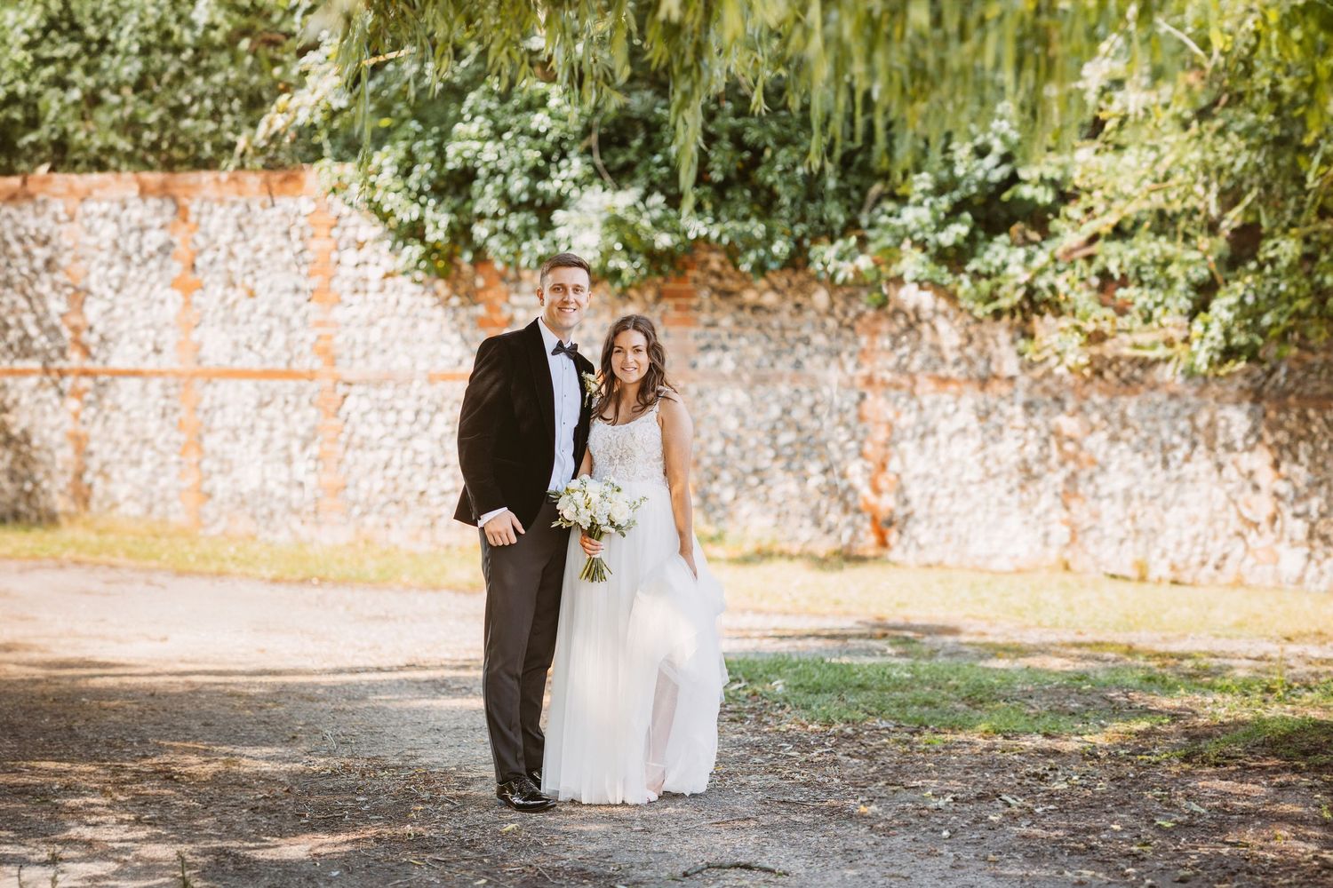 Couple poses for portrait against weathered brick wall in garden setting.
