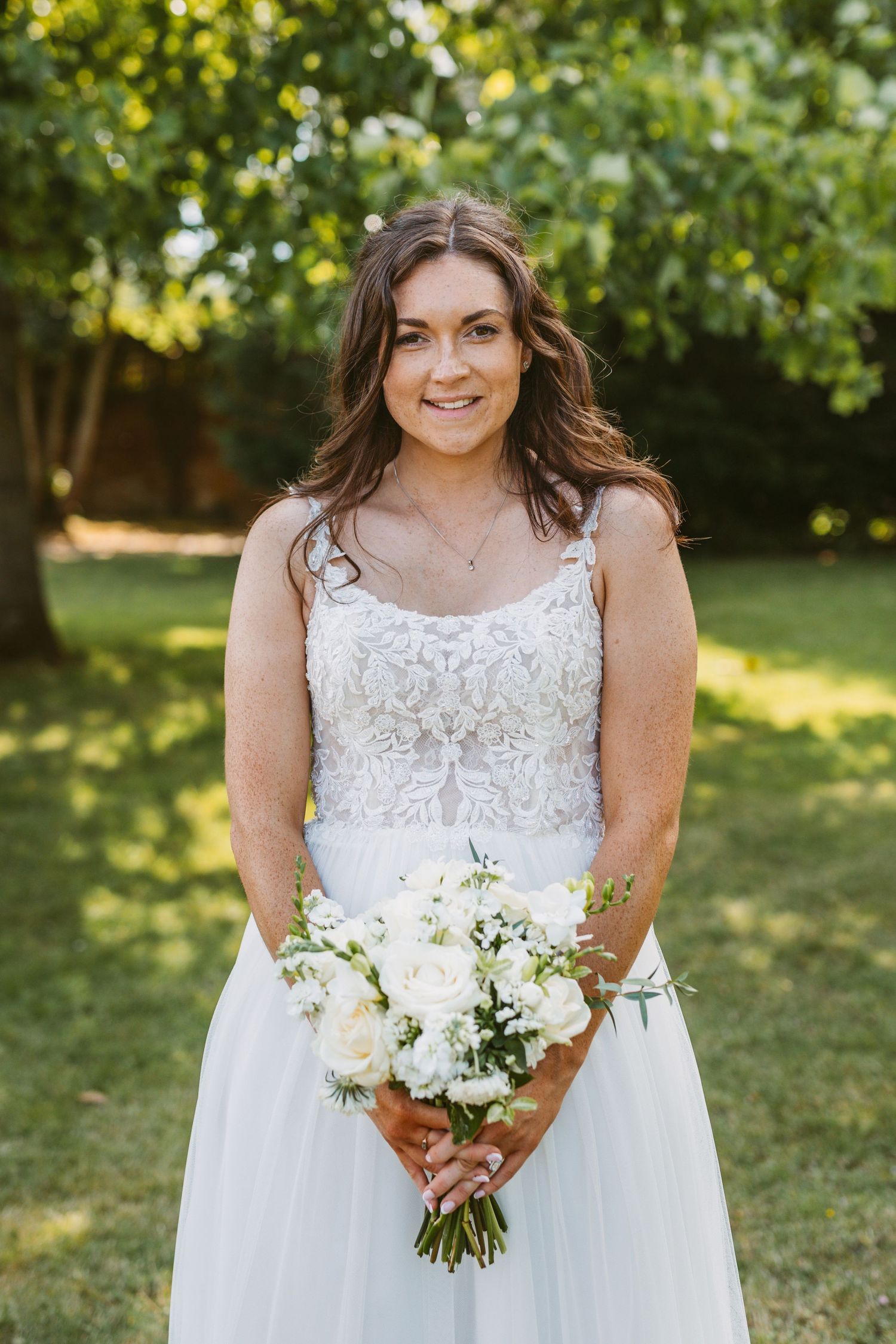 Person in lace wedding gown holds white floral bouquet while smiling in garden setting.
