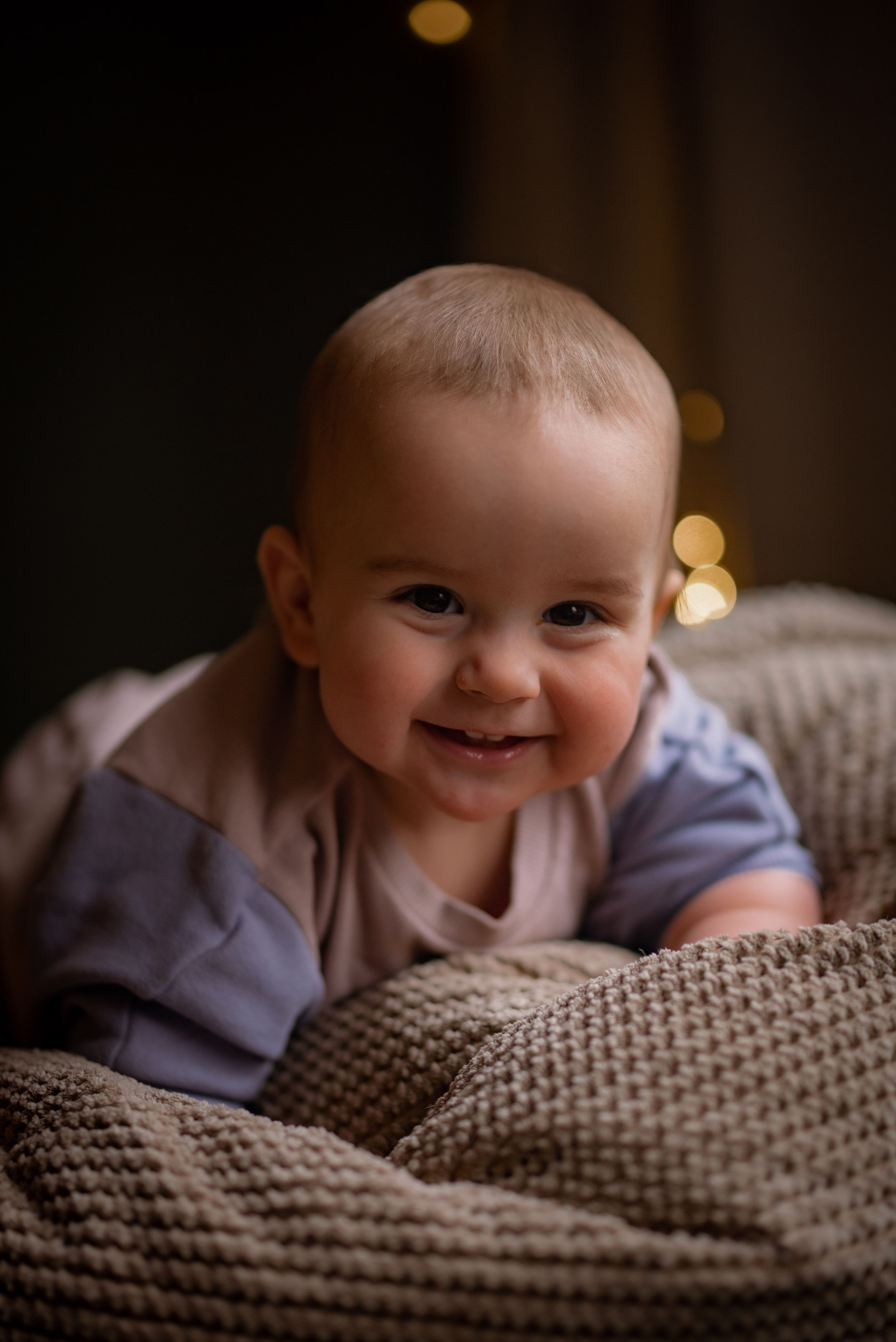 baby in light purple and cream leans forward on a beige beanbag smiling at the camera