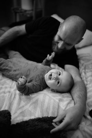 dad and baby lie on bed together. the baby looks at the camera smiling