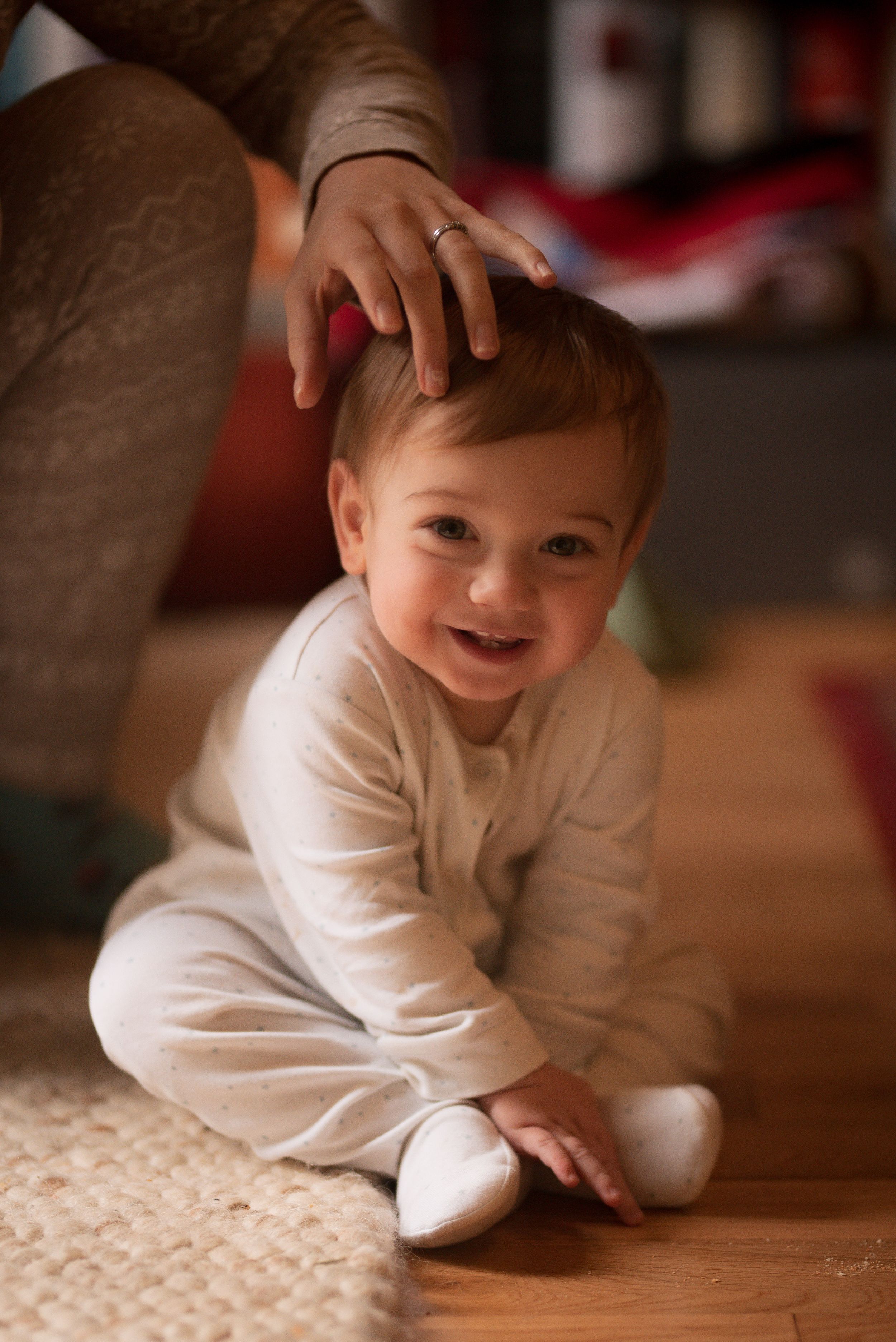 little boy with light brown hair in white sits cross legged and smiles at the camera