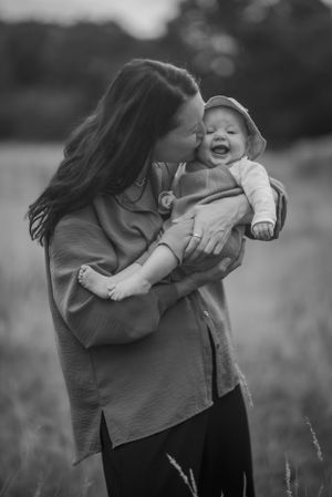 mum with long dark hair cradles her baby and kisses her cheek while stood in long grass