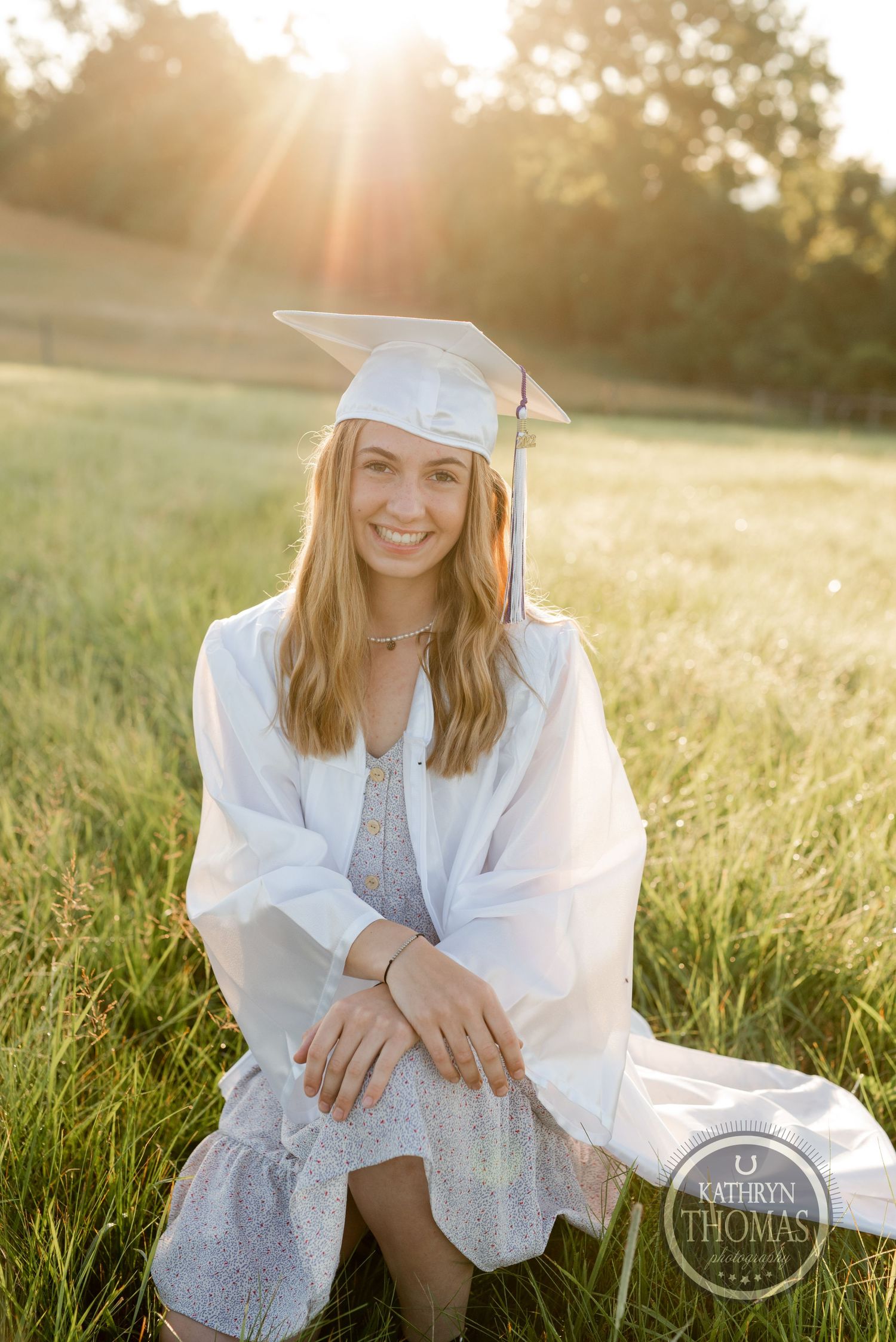 Sunset graduation portrait in pastoral field with graduate in white cap and gown.