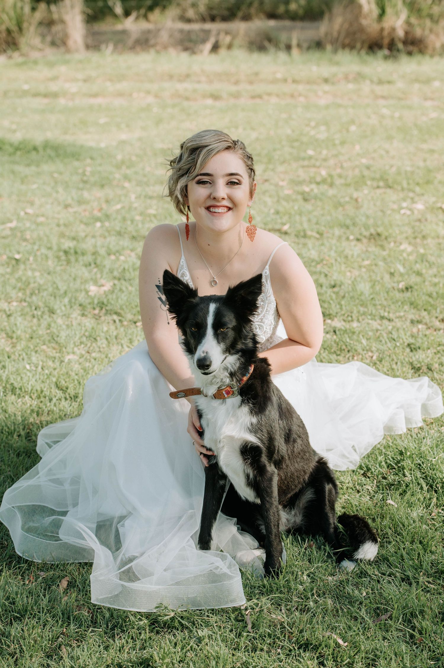 A bride in a white dress poses with a black and white border collie dog on a grassy field.