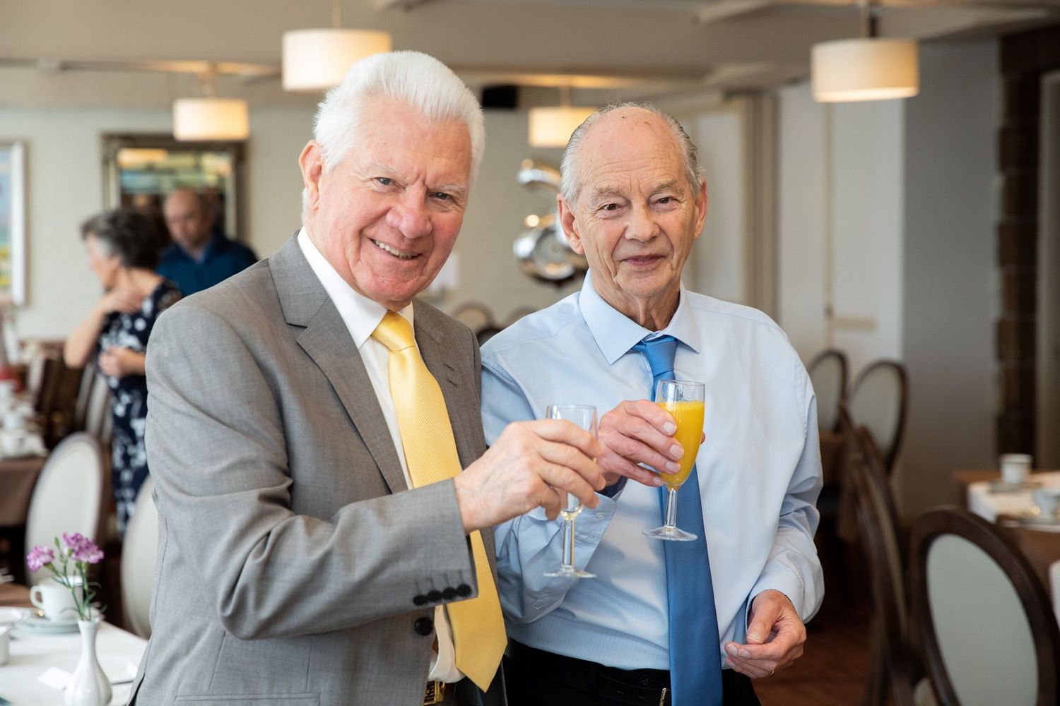Two seniors in formal attire sharing drinks and laughing at an elegant indoor celebration.