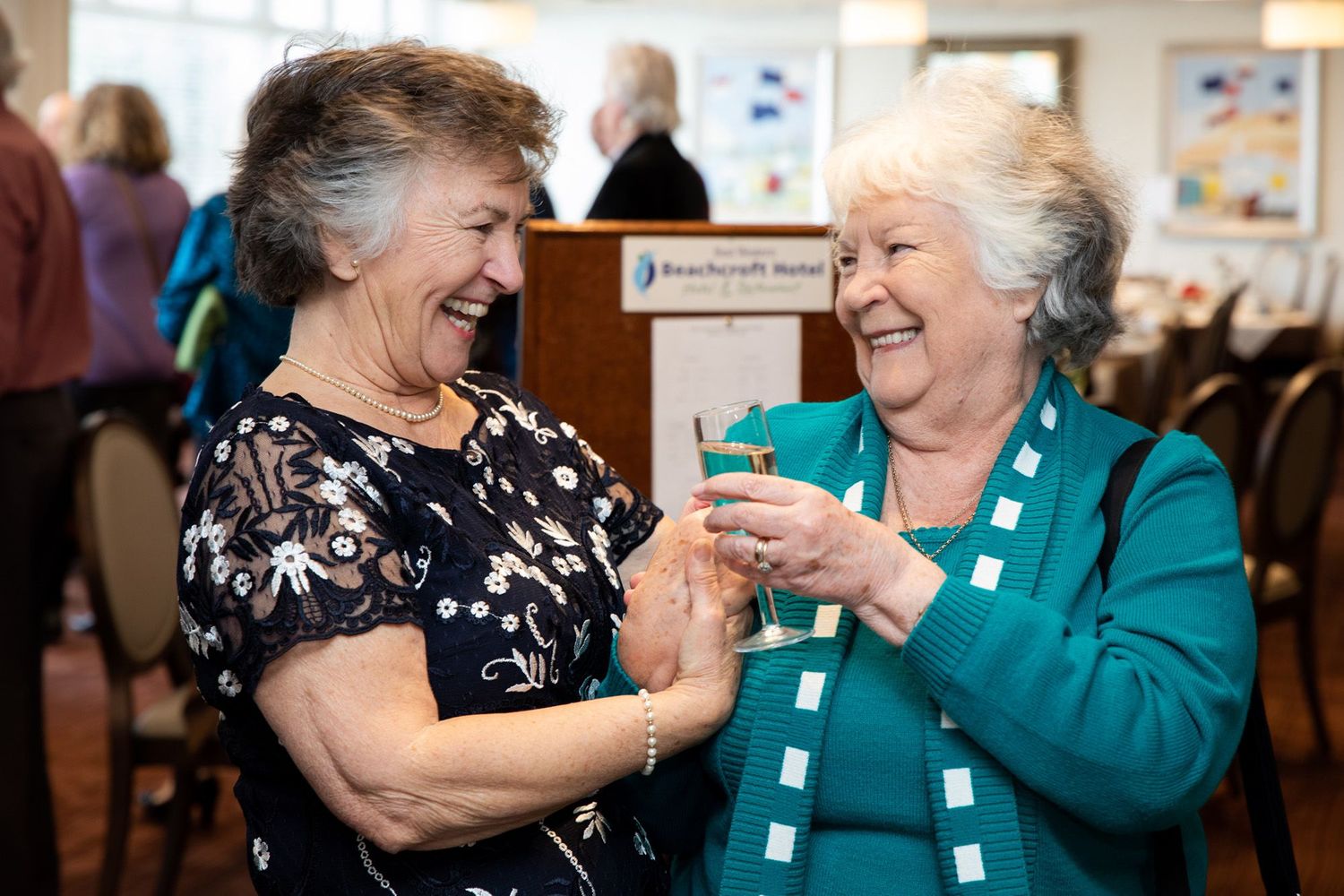 Two senior ladies sharing a joyful moment while holding drinks at a social gathering.