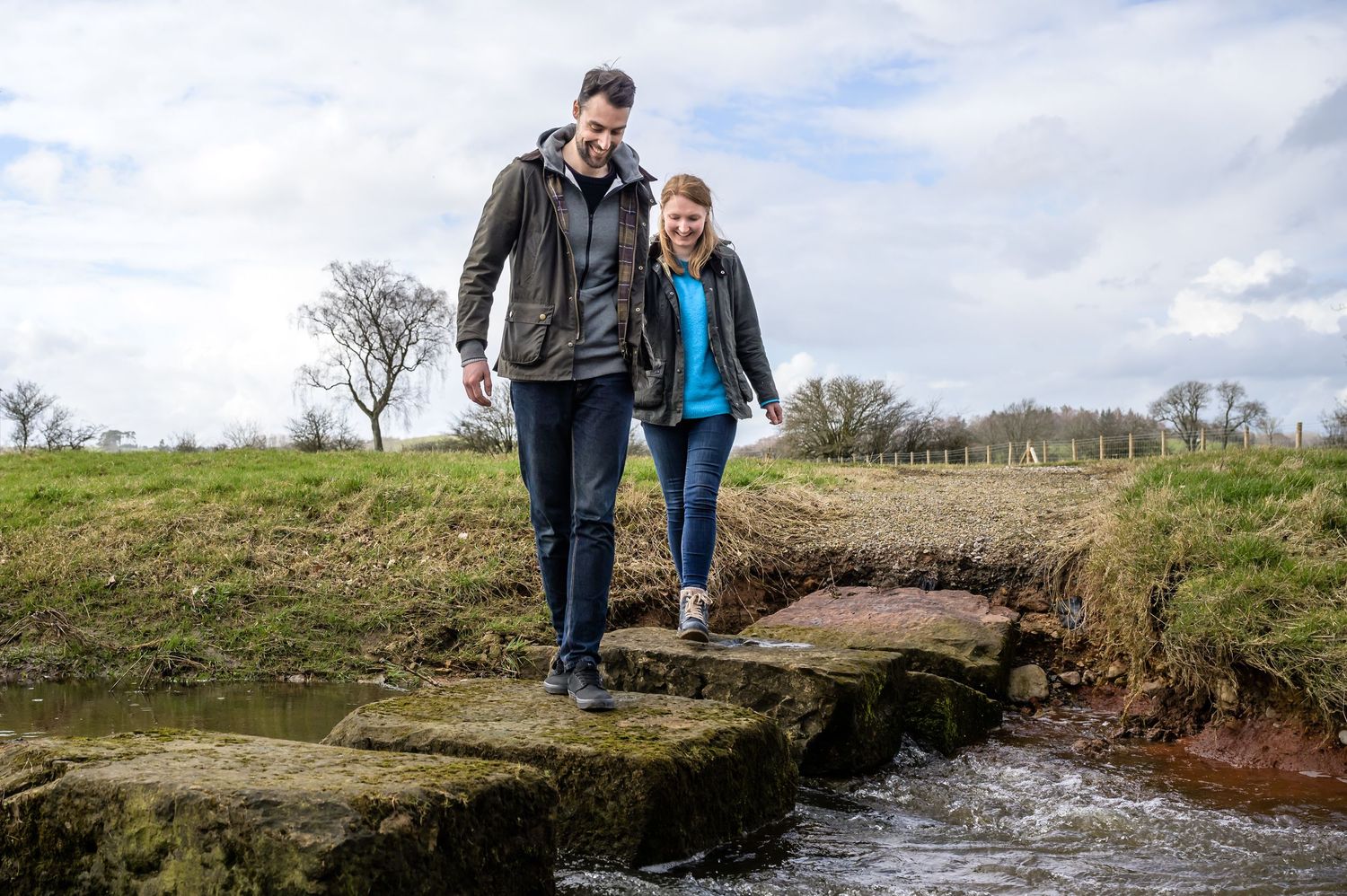 Engaged couple walk over stepping stones at Eden Barn in Lake District