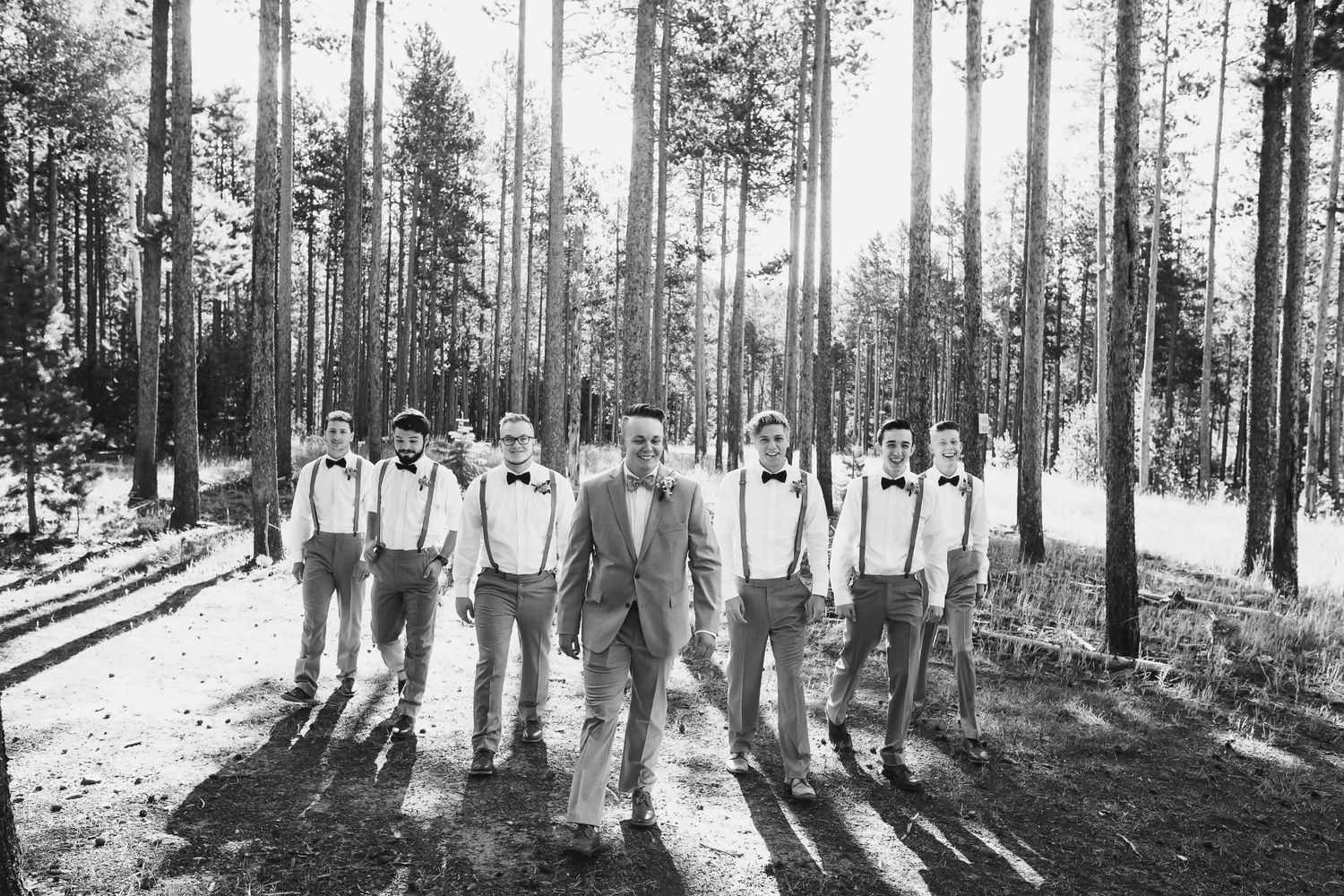 A black and white photo of a wedding party in suspenders standing among tall pine trees.