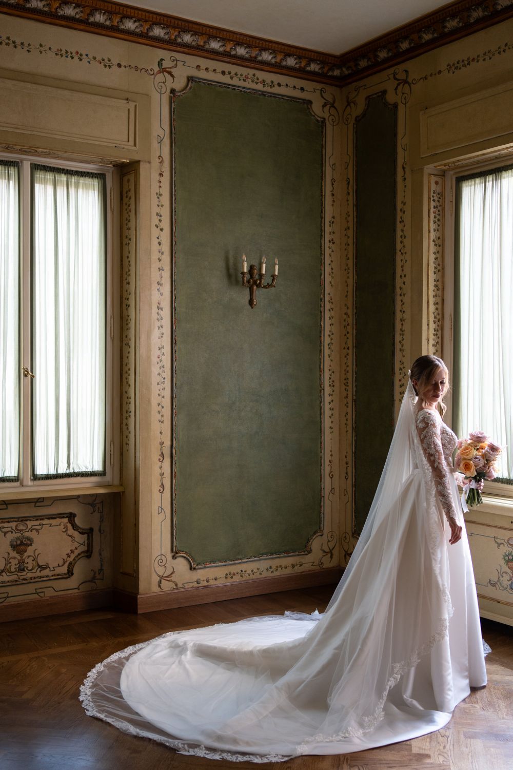 Bride in flowing white gown with long train poses in ornate room with green walls.