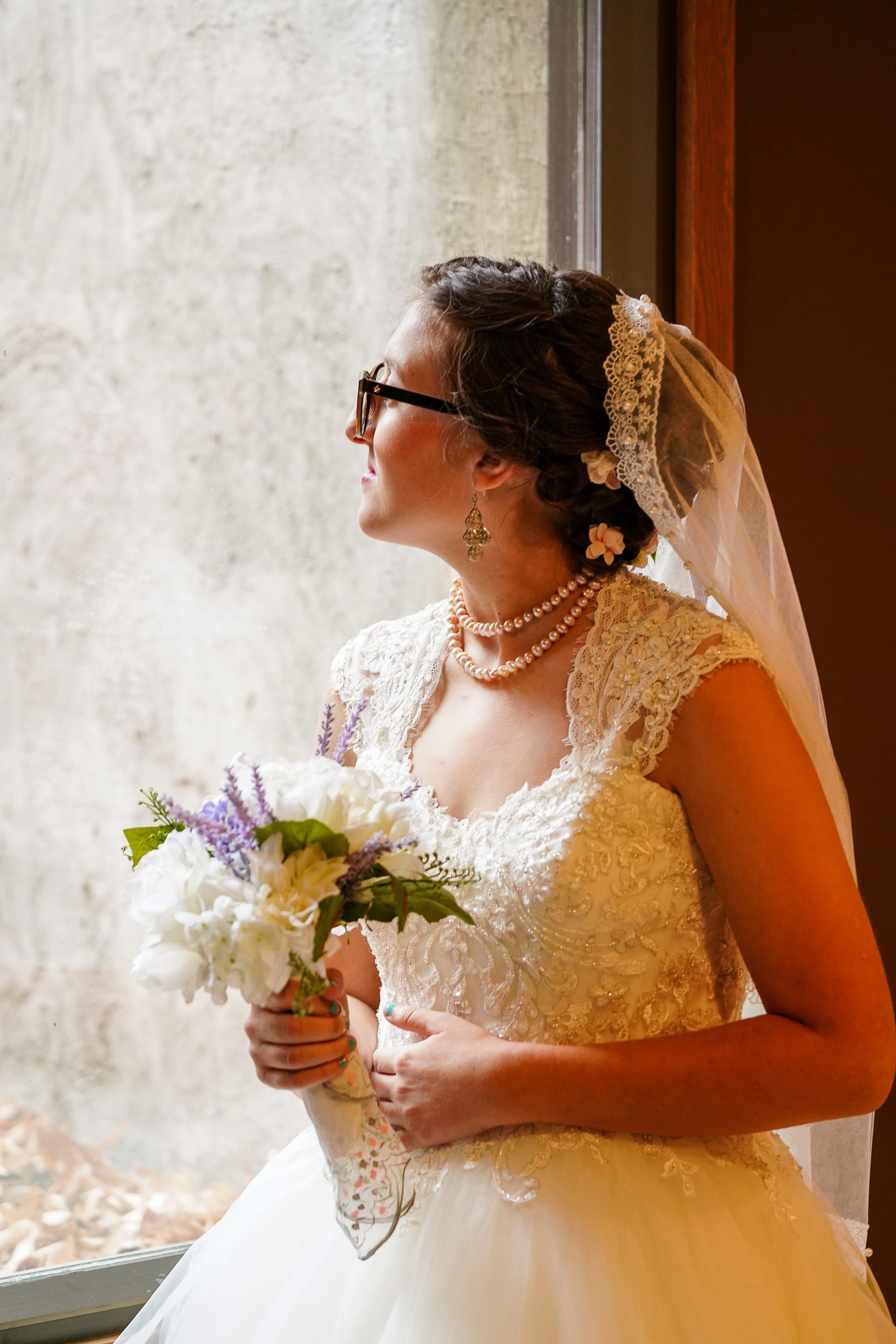 A bride in a lace wedding dress holds a bouquet while looking out a window.
