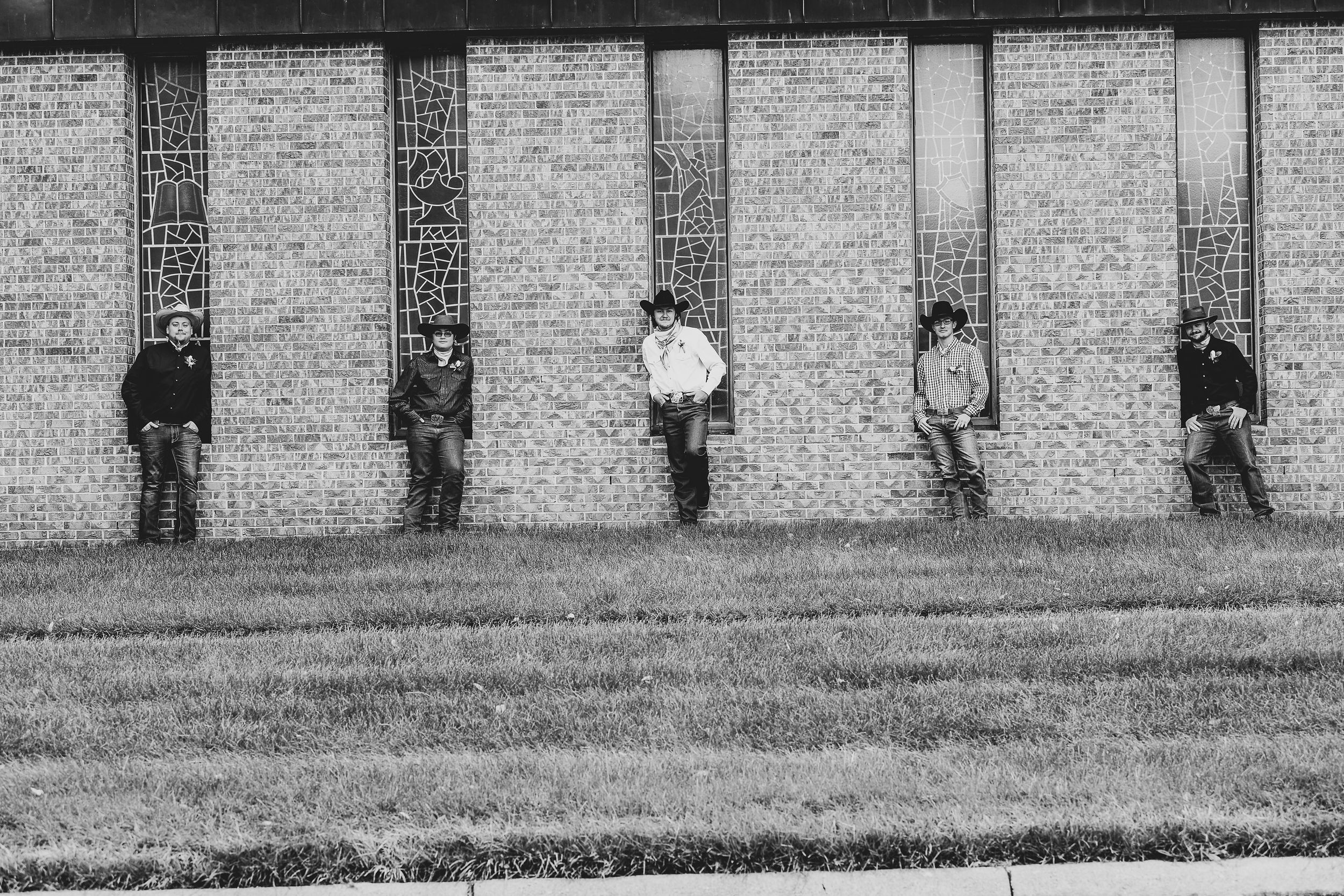 Five people lean against a brick wall in a black and white artistic composition.