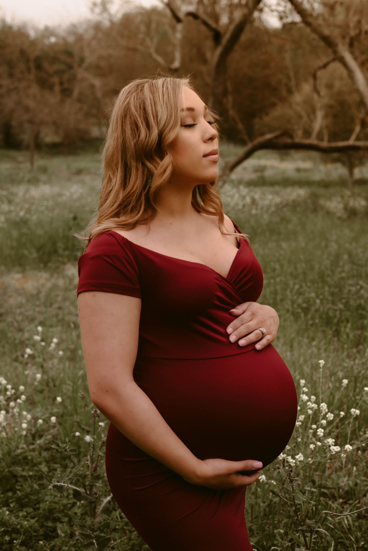 Dreamy side profile maternity photo featuring a burgundy dress against a natural outdoor backdrop.