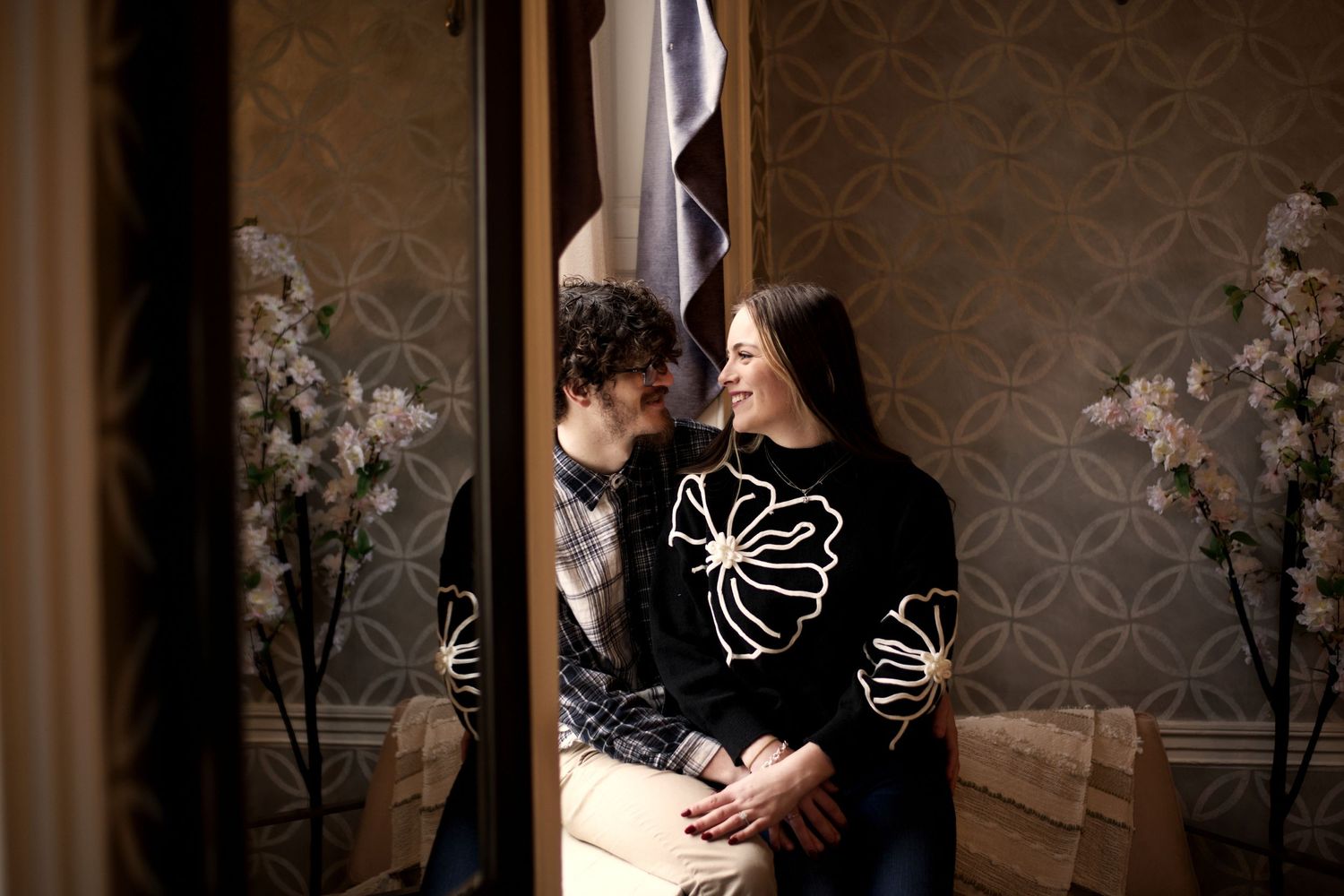 A couple in matching black sweaters embraces in a dimly lit room with floral decorations.