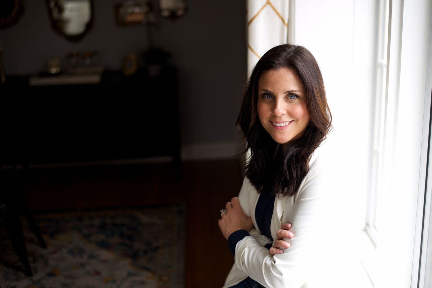 Portrait of someone with dark hair and a white sweater smiling by a bright window in a cozy home setting.