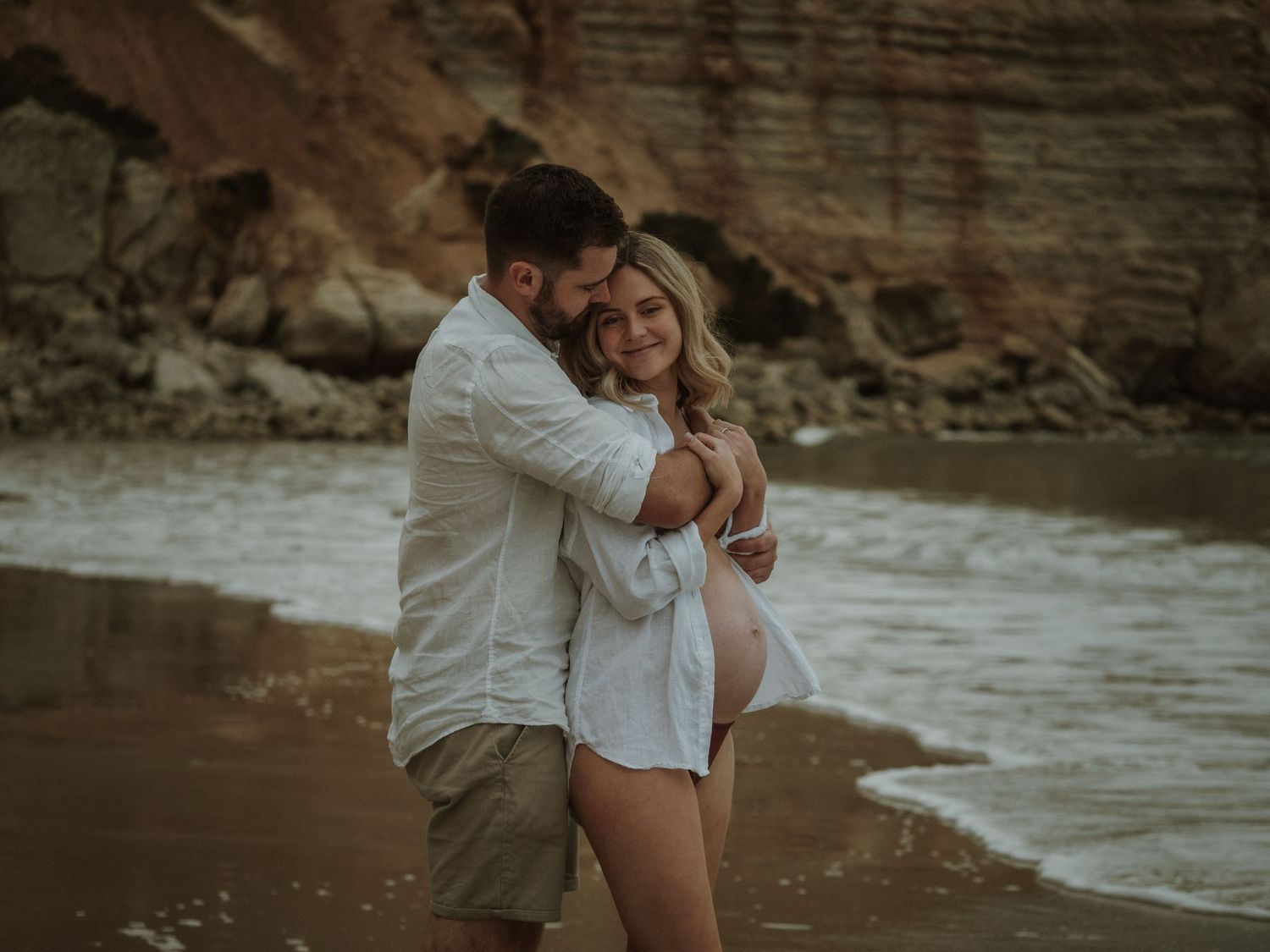 A tender embrace between a couple on a beach with red rock cliffs in the background.