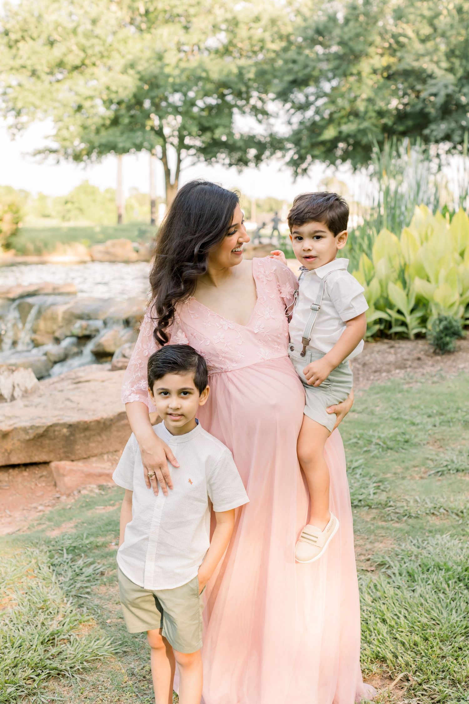 A woman in a flowing pink dress poses with two children outdoors in a natural setting.