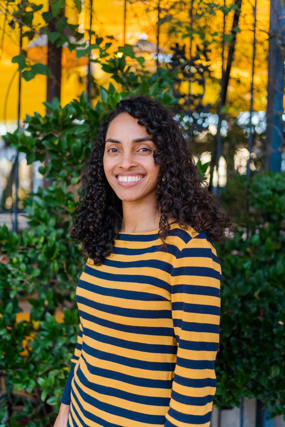 Cheerful person wearing yellow and navy striped long sleeve top poses outdoors against greenery and yellow wall.