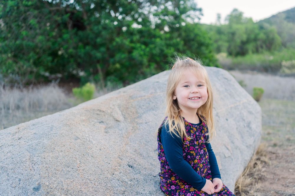 Child wearing navy and floral dress sits on large boulder in outdoor nature setting with green foliage.