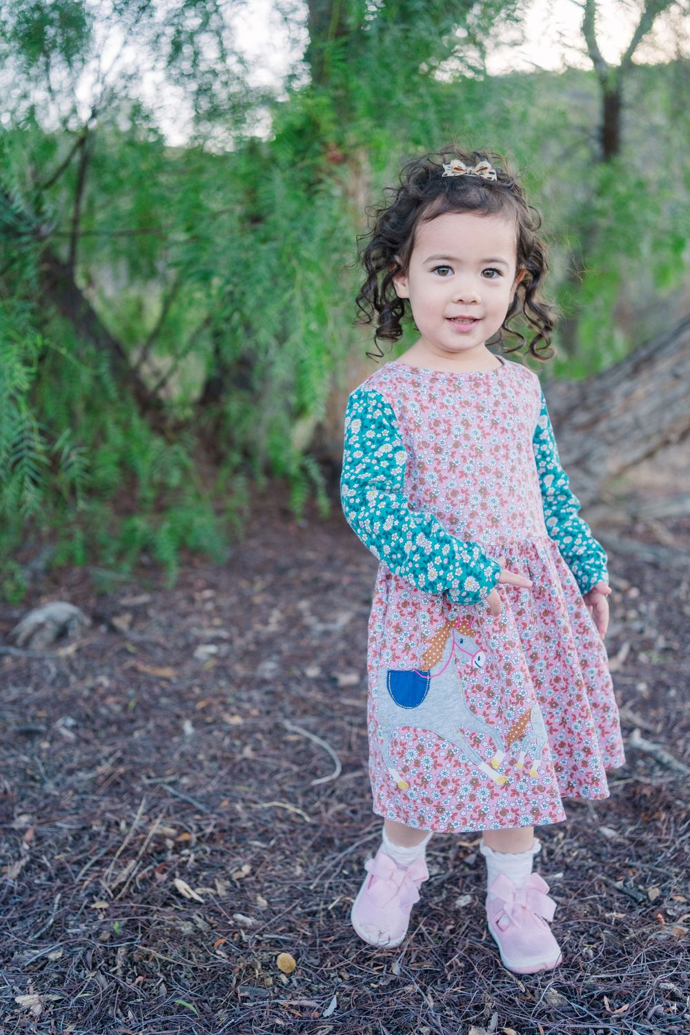 Young child wearing a floral print dress with contrasting sleeves stands outdoors among green foliage.