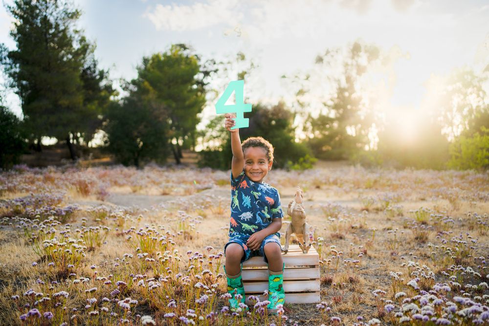 A child sits on stacked books outdoors wearing a blue sweater and party hat during golden hour sunset.