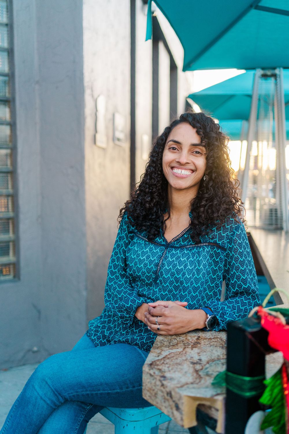 A person with curly hair wearing a blue patterned top and jeans sits outdoors under turquoise umbrellas.