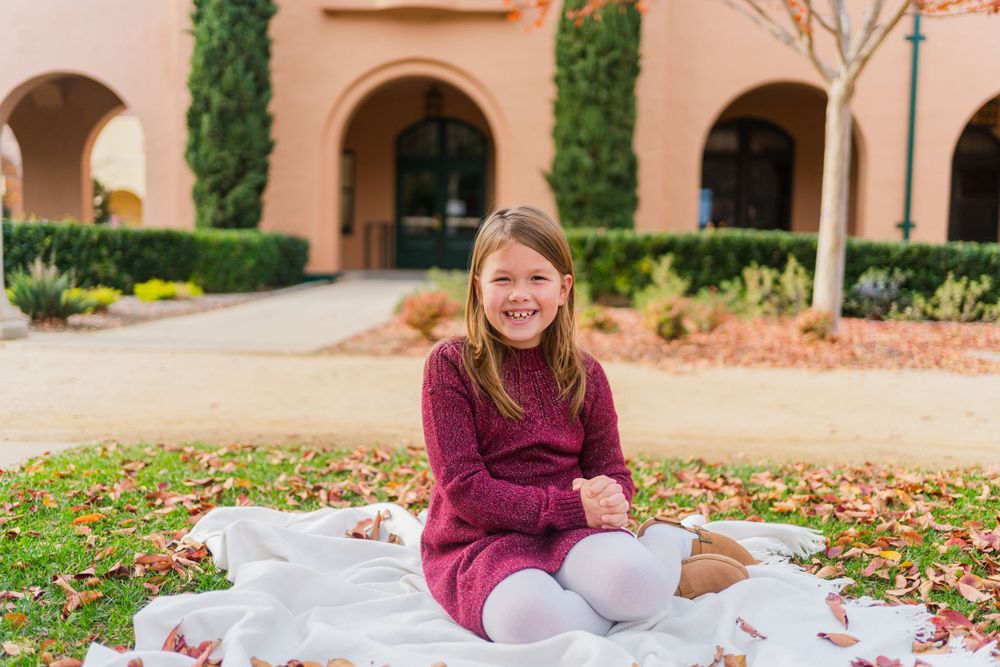 A child in a burgundy sweater dress sits on a white blanket among fall leaves near a Mediterranean style building.