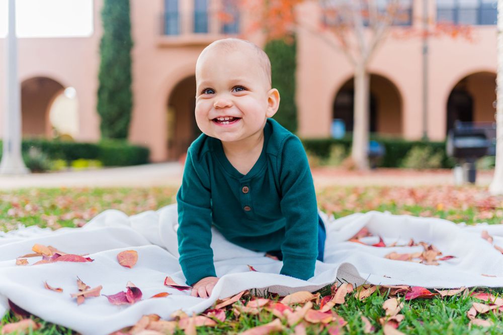 A cheerful baby in a teal sweater sits on a white blanket surrounded by autumn leaves on the ground.