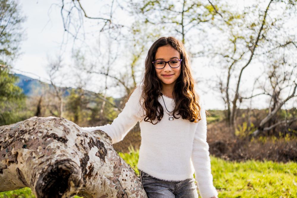 A student with glasses and wavy dark hair wears a white sweater while posing near a large rock in a sunny outdoor setting.