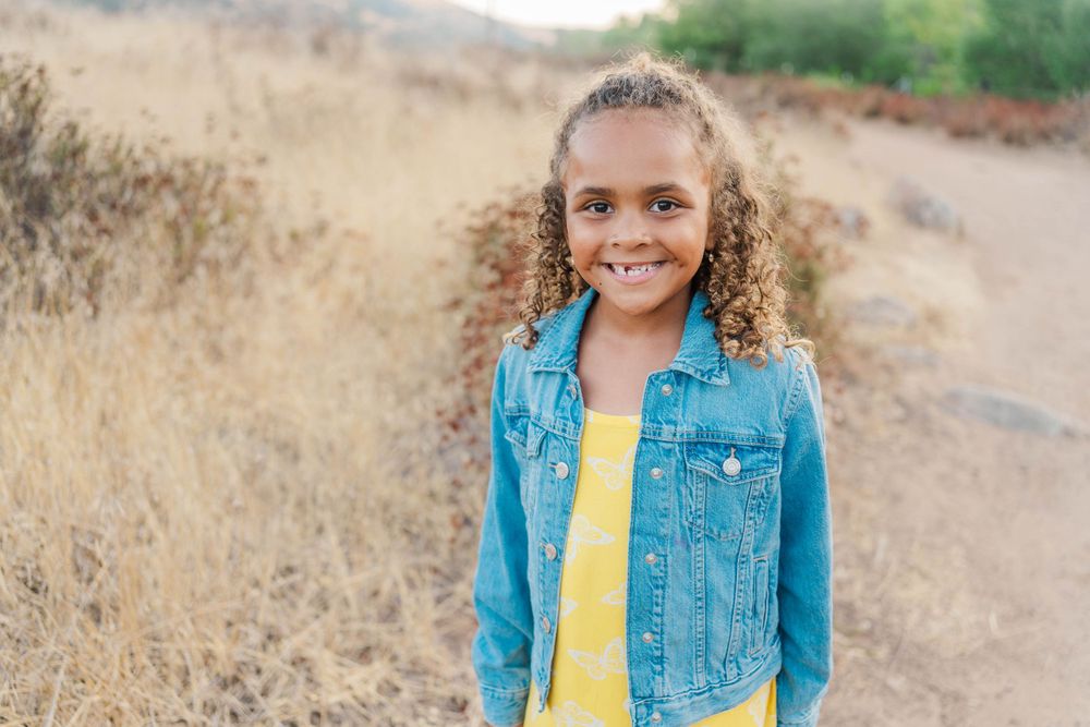 A child with curly hair wearing a denim jacket and yellow shirt stands on a dirt path among dry grass.