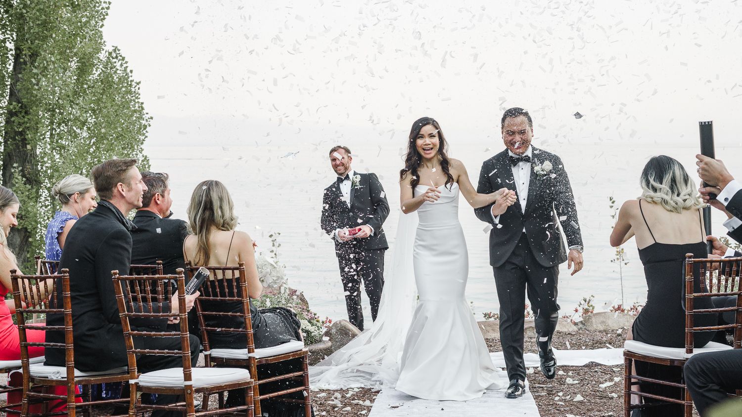 A celebratory moment at an elegant outdoor wedding with falling confetti and wooden chiavari chairs along the aisle.