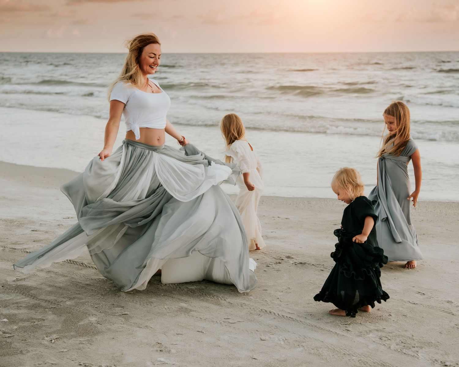 A mother and three daughters play on a sandy beach at sunset, wearing flowing gray and black dresses.