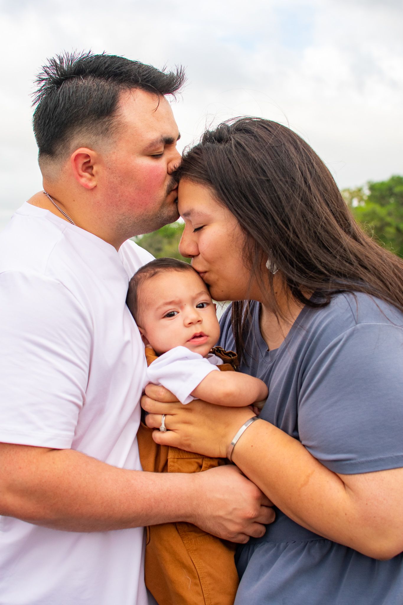 Intimate family moment captured outdoors with baby in brown overalls.