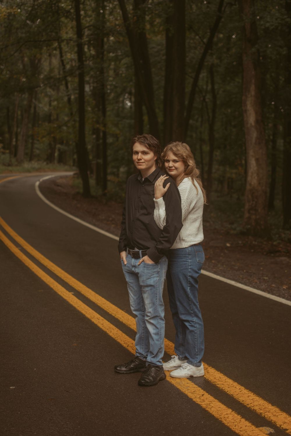 A couple stands together on yellow road lines through a wooded area.