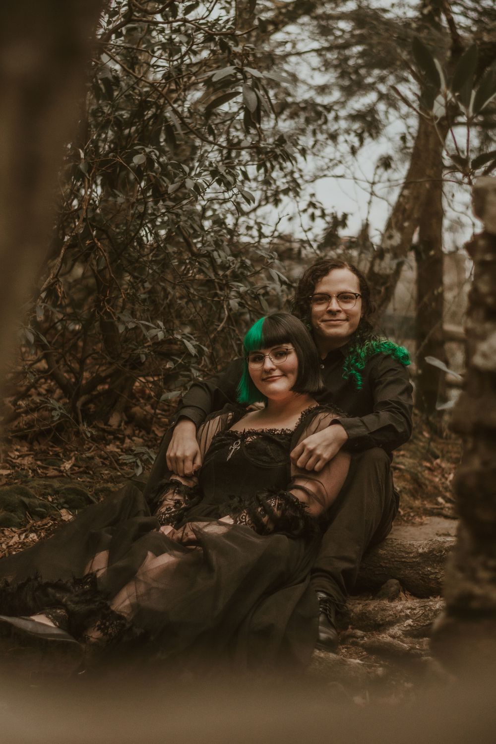 Couple in dark clothing sharing intimate moment while sitting among forest trees.