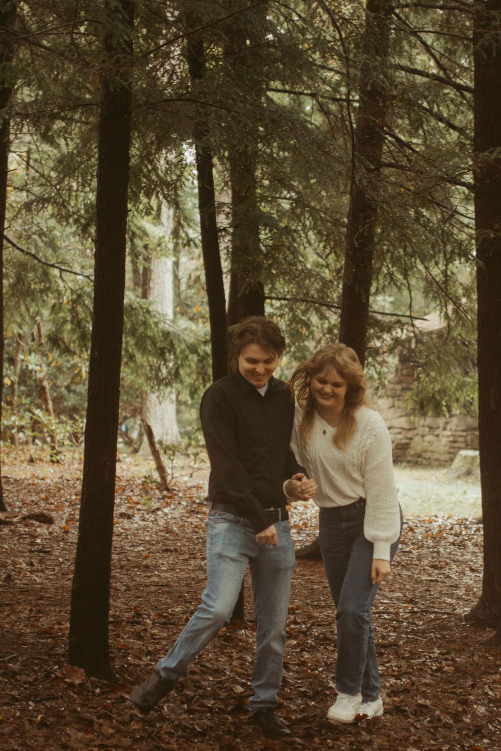 A couple laughs together while walking through a wooded forest trail with fallen leaves.