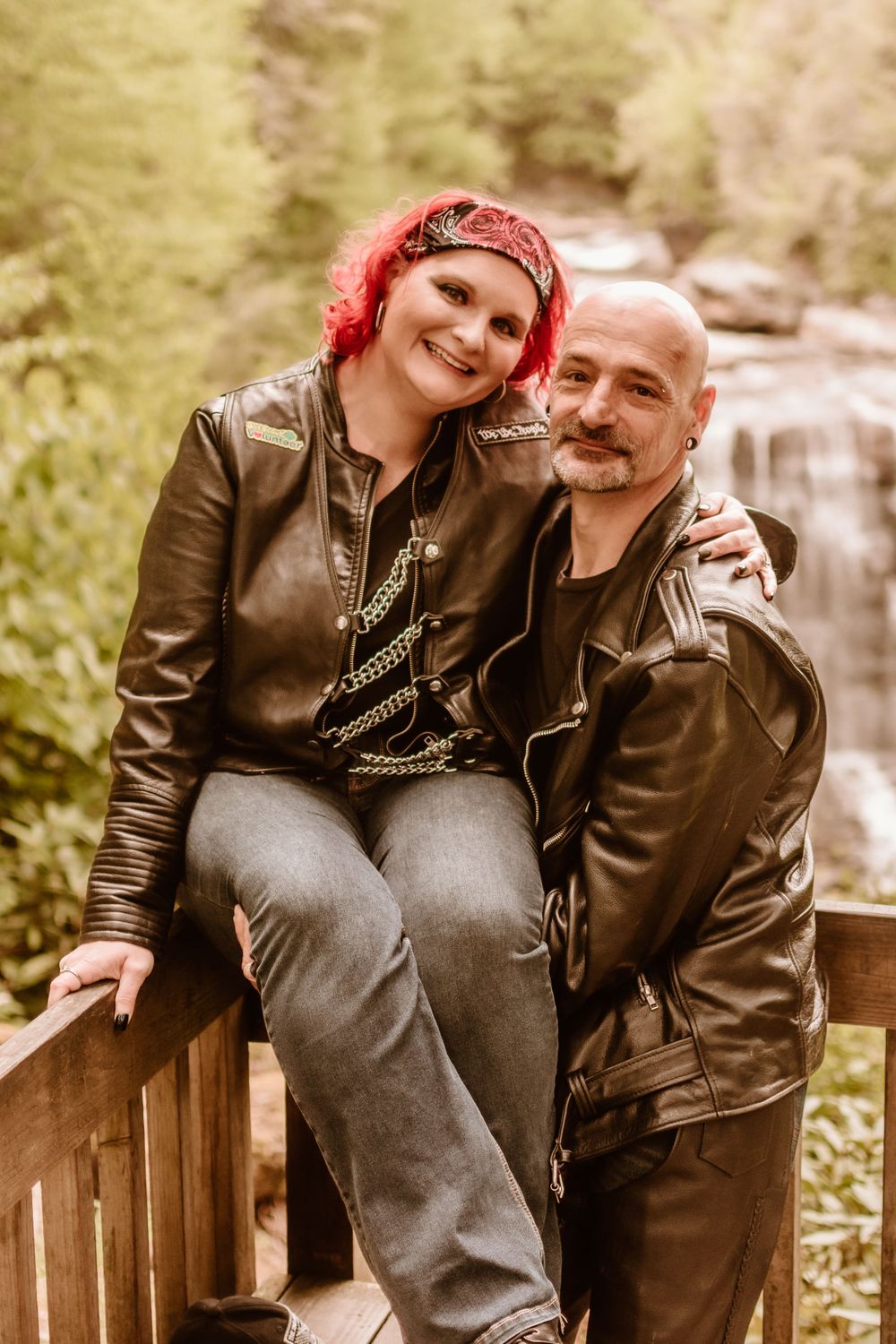 Couple in leather jackets sharing an intimate moment on a wooden deck with waterfall backdrop.