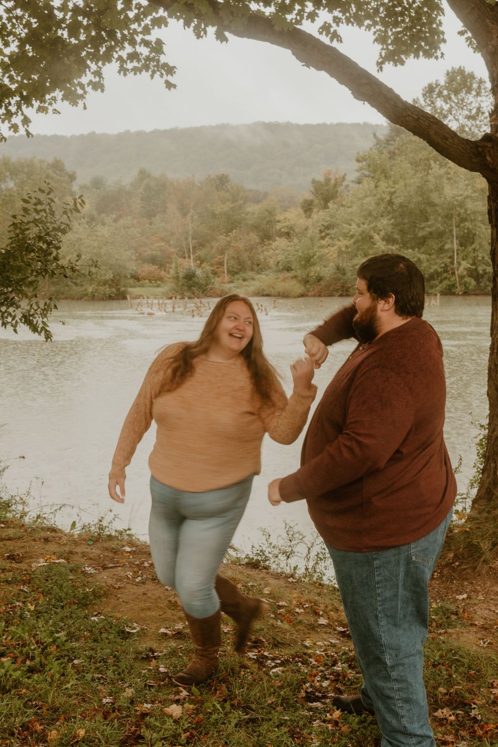 A playful couple shares a laugh by a scenic lake surrounded by autumn trees.