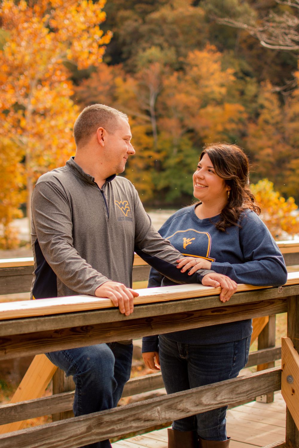 A couple shares a tender moment on a wooden bridge with fall colors in background.